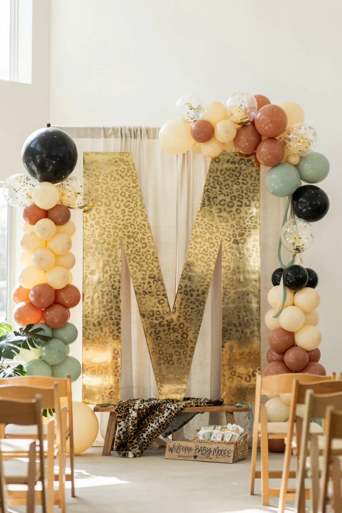 A statement wall with a giant number (month of baby) or oversized letter (baby's initial) in gold or animal print, surrounded by safari-colored confetti balloons. Guests stand in front for photos.