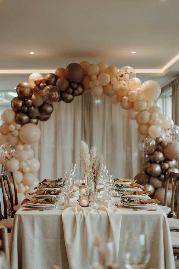 Elegant balloon garland in gold, khaki, and cream tones forming an arch over a styled table. Mixed matte and metallic finishes.