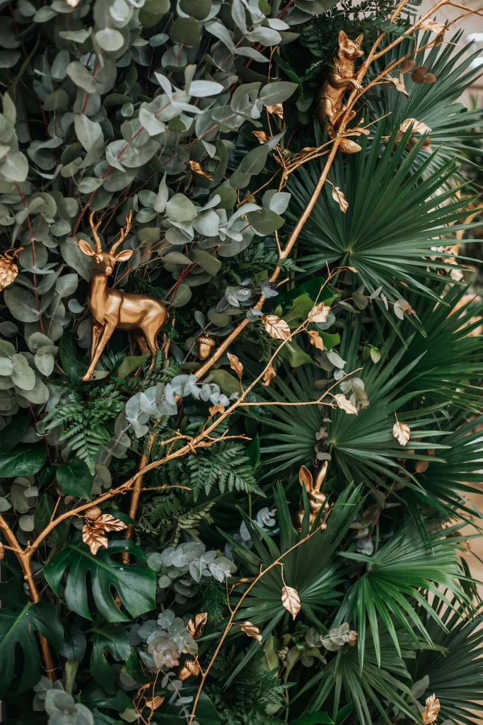 Dense wall of faux eucalyptus, palm fronds, and jungle greenery with scattered gold-painted branches and small gold animal figurines nestled in the foliage. Warm lighting.
