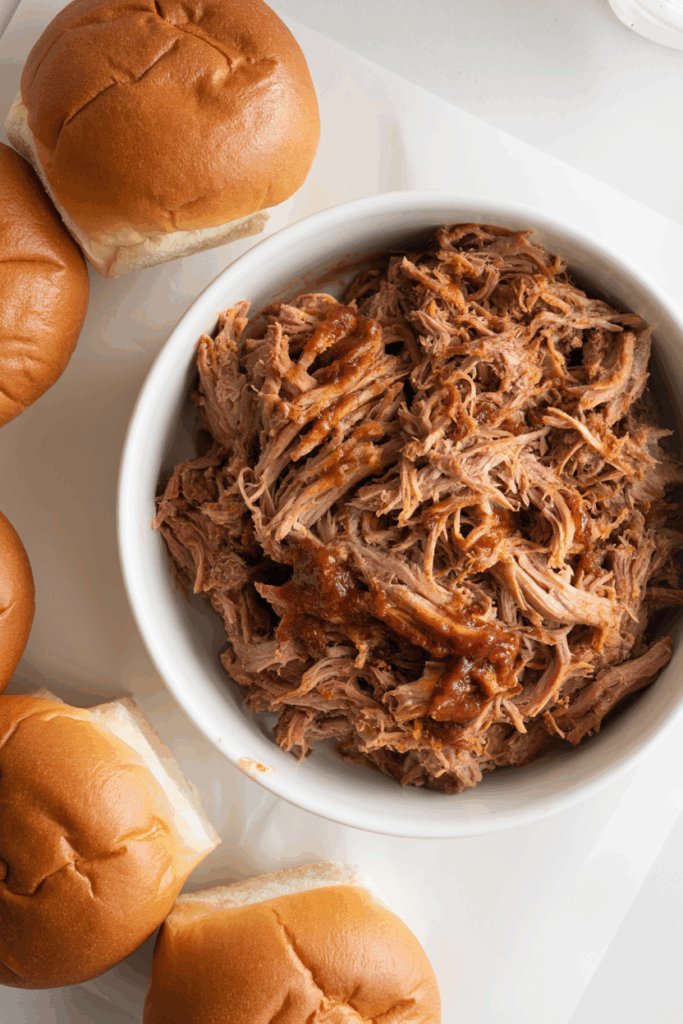 BBQ pulled pork piled in a white dish with brioche buns in the background under warm diffused lighting.