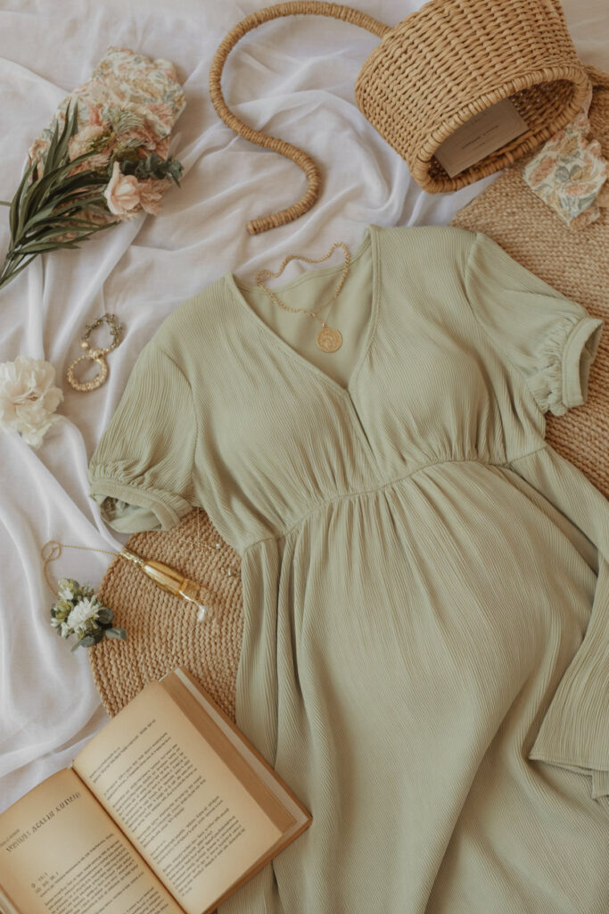 Flatlay of soft pastel dresses, sage maternity dress, woven accessories, and gold jewelry arranged for outfit inspiration.