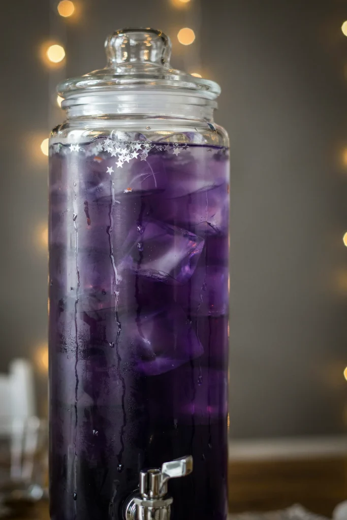A glass beverage dispenser filled with a deep purple-blue layered punch, garnished with edible silver stars floating on top, condensation on the glass.