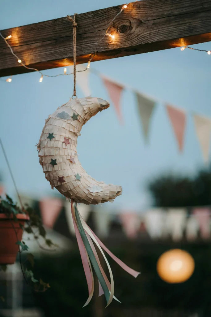 A large crescent moon pull-string piñata hanging from a doorway or ceiling beam, styled with trailing ribbon and surrounded by string lights.