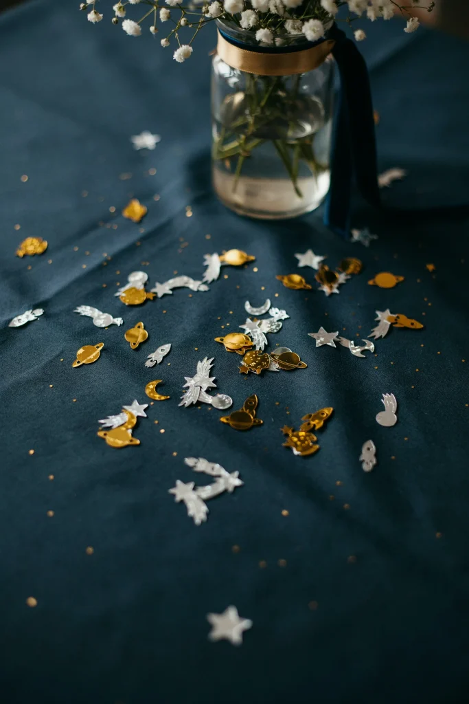 A close-up of a navy tablecloth scattered with gold and silver space-shaped confetti — tiny planets, stars, and rocket shapes — beneath a centerpiece arrangement.