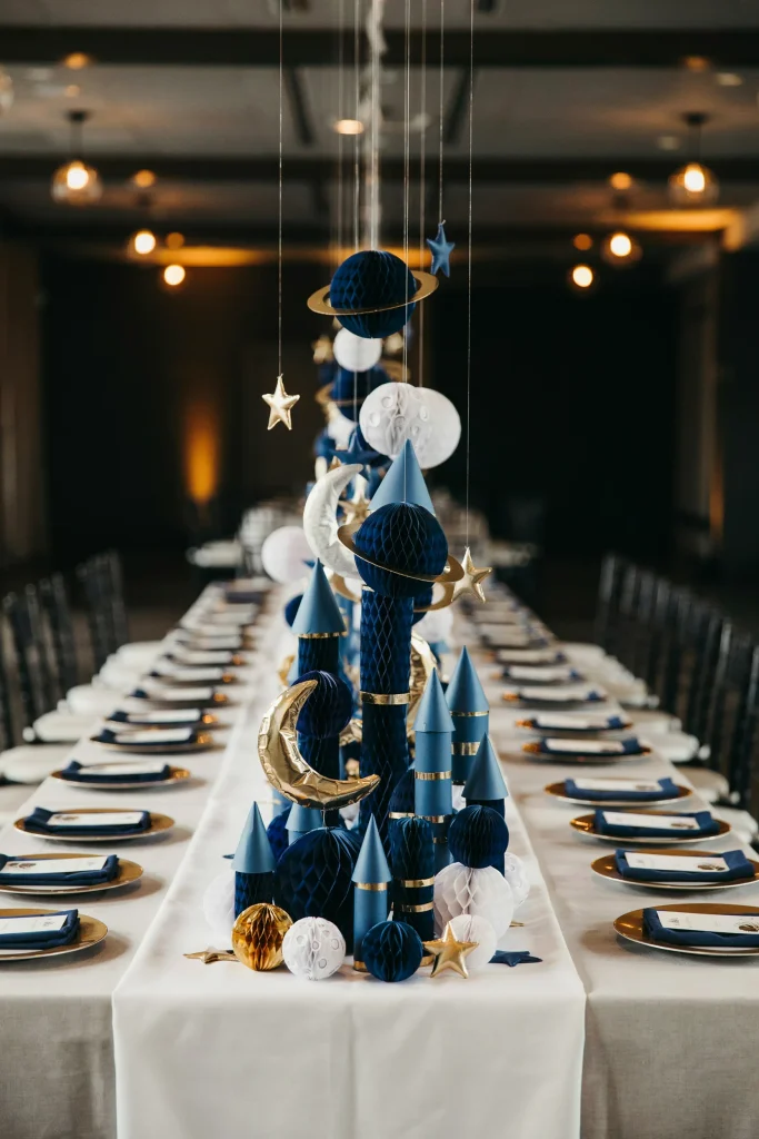 A table covered in layered honeycomb rockets, planets, moons, and stars in navy, white, and metallic gold, clustered together at varying heights down the center of a long table.