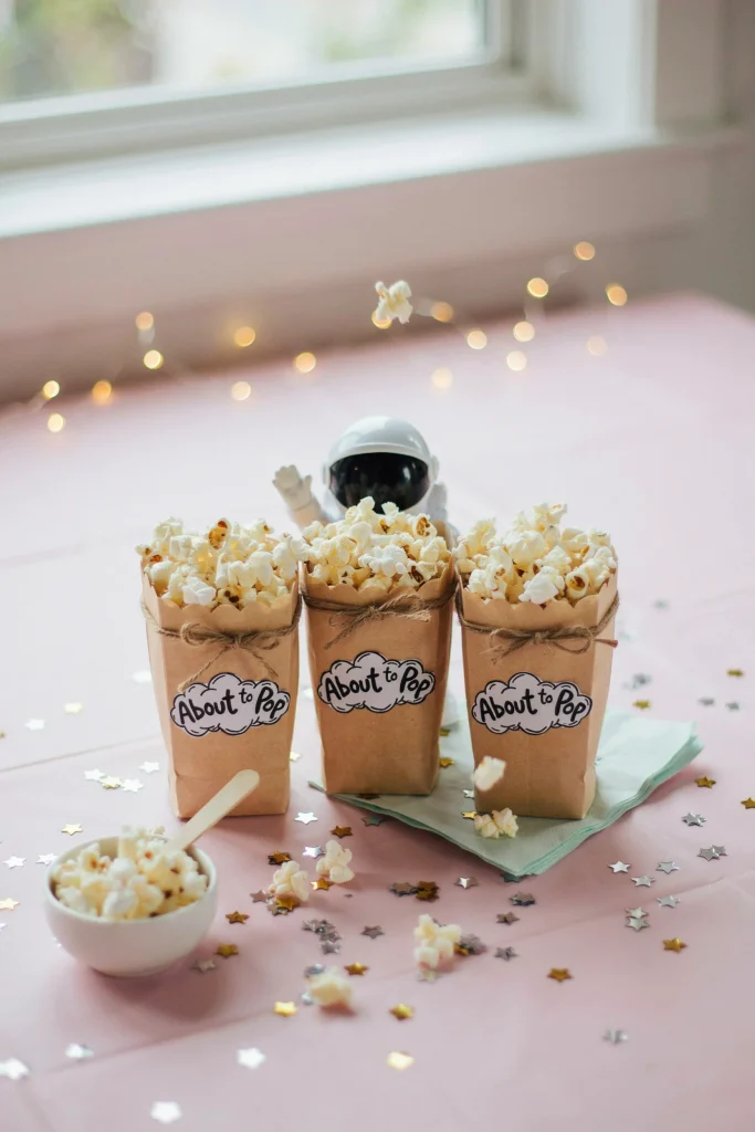 A small dessert station with popcorn bags labeled "About to Pop," styled with star confetti and a small astronaut figurine peeking out from behind the bags.