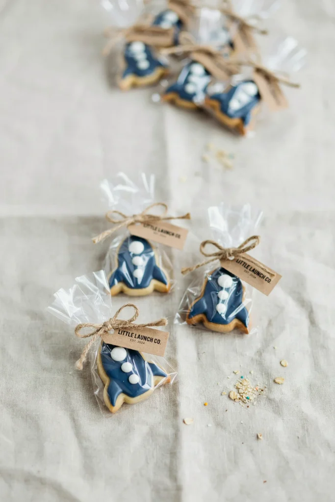 Individually packaged sugar cookies decorated as simple rocket ships in royal icing, navy and white, tied with twine and a small tag.