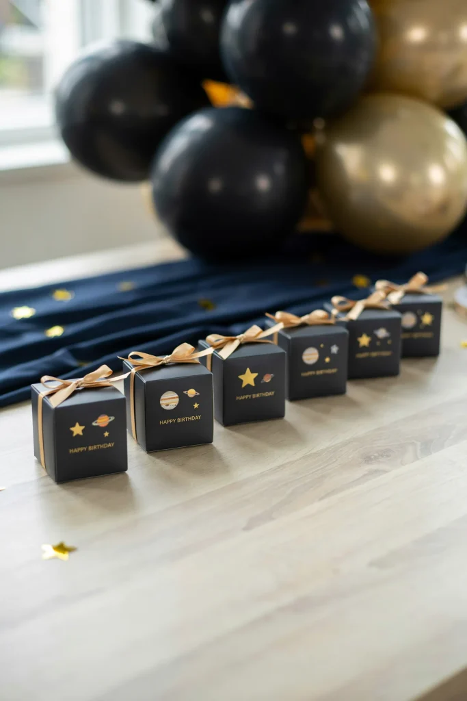 A row of small black favor boxes with gold star and planet stickers, lined up on a table, each tied with a simple ribbon.