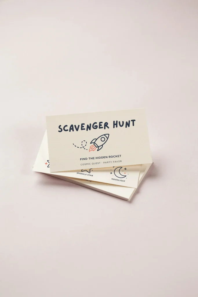 A small stack of printed scavenger hunt cards with space-themed icons — a hidden rocket, a star, a moon — designed for guests to find items around the party space.