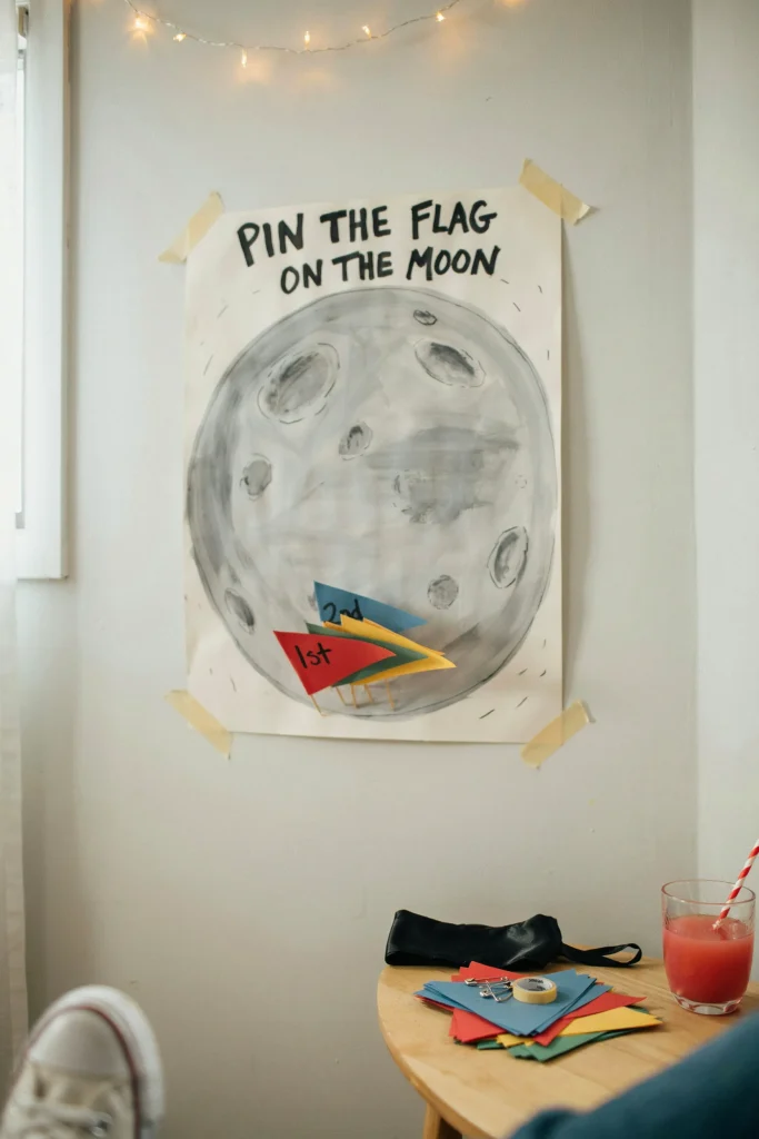 A large poster-board moon surface taped to a wall, with small paper flags and a blindfold resting on a nearby table, styled like a classic party game corner.