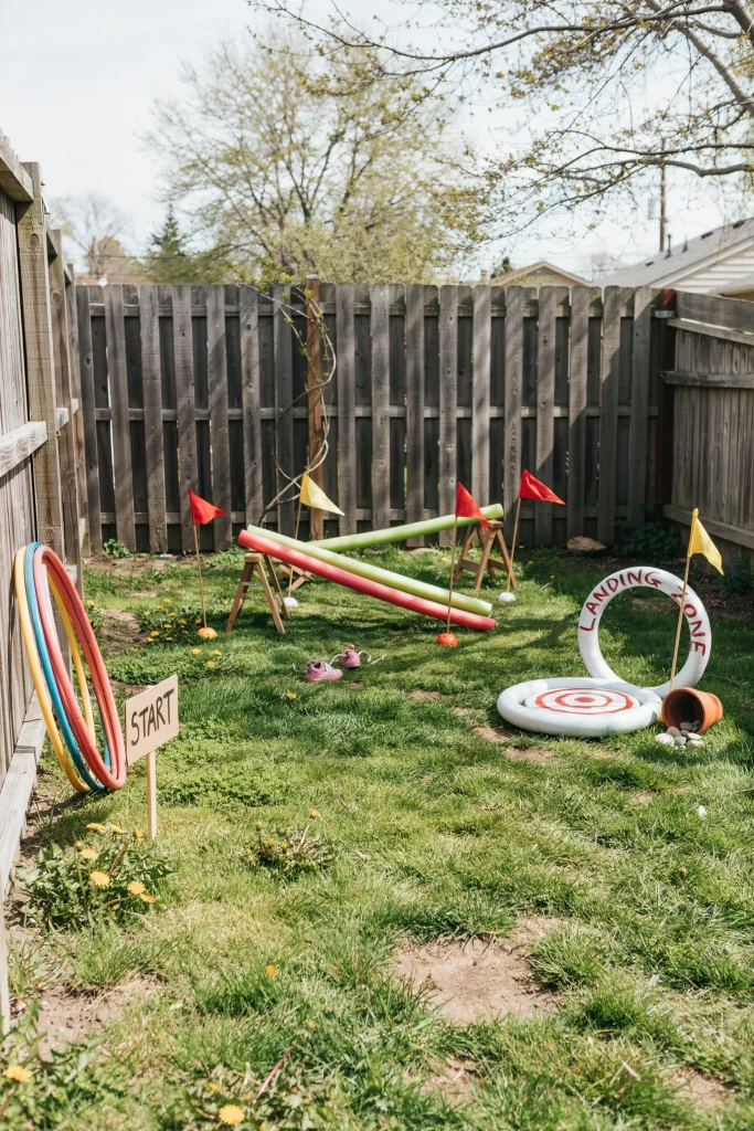 A small backyard setup with hula hoops, a balance beam made of pool noodles, and a "landing pad" hula hoop target, styled with star flags marking each station.