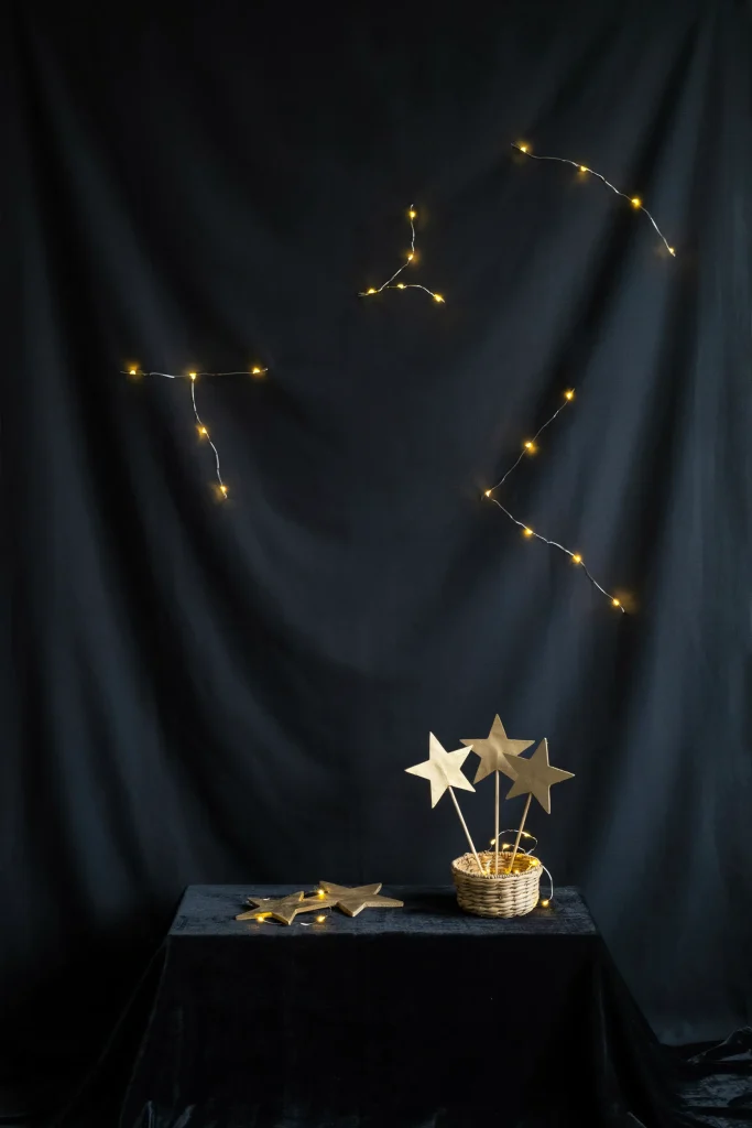 A black backdrop with small string lights arranged to mimic real constellations, with simple gold star props on sticks resting nearby for guests to hold.