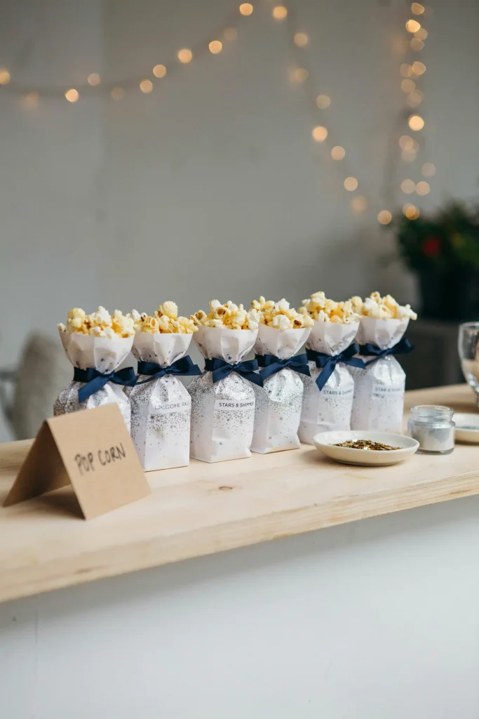 Small white paper bags of popcorn dusted with edible silver shimmer and tiny star sprinkles, tied with navy ribbon, lined up as a grab-and-go snack station.