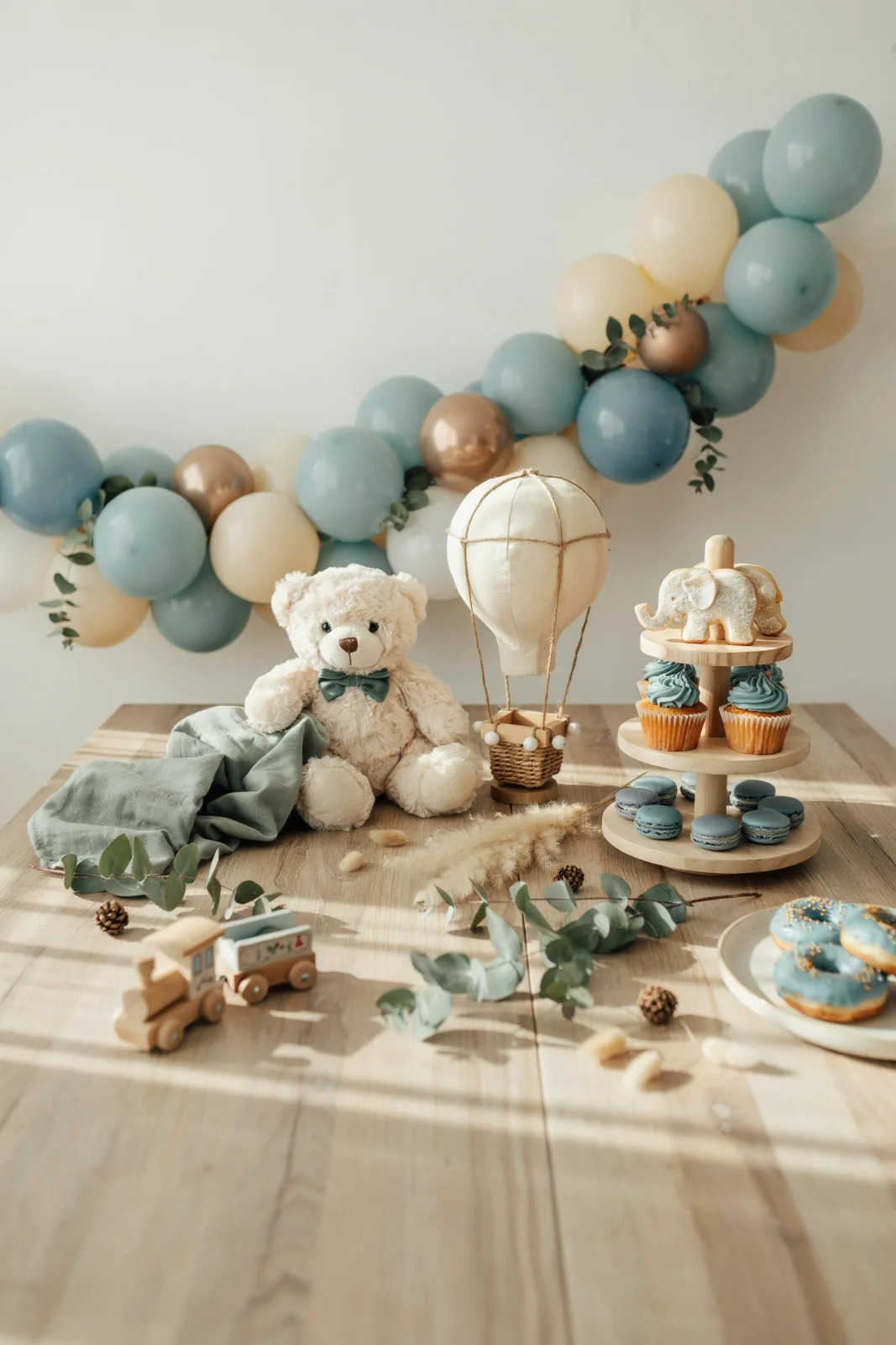 A flat-lay of a beautifully styled boy baby shower dessert table featuring a soft dusty-blue and sage color palette, balloon garland arch in the background, wooden "It's a Boy" letters as the centerpiece, warm natural light,