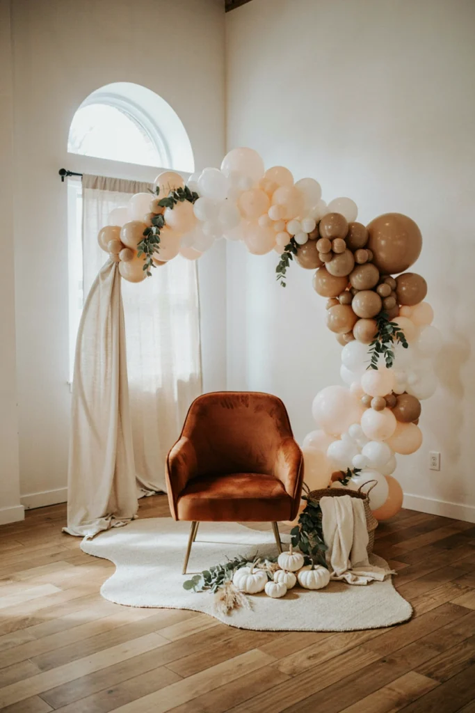 A balloon arch in champagne, ivory, and blush tones framing a seating area with a single velvet accent chair