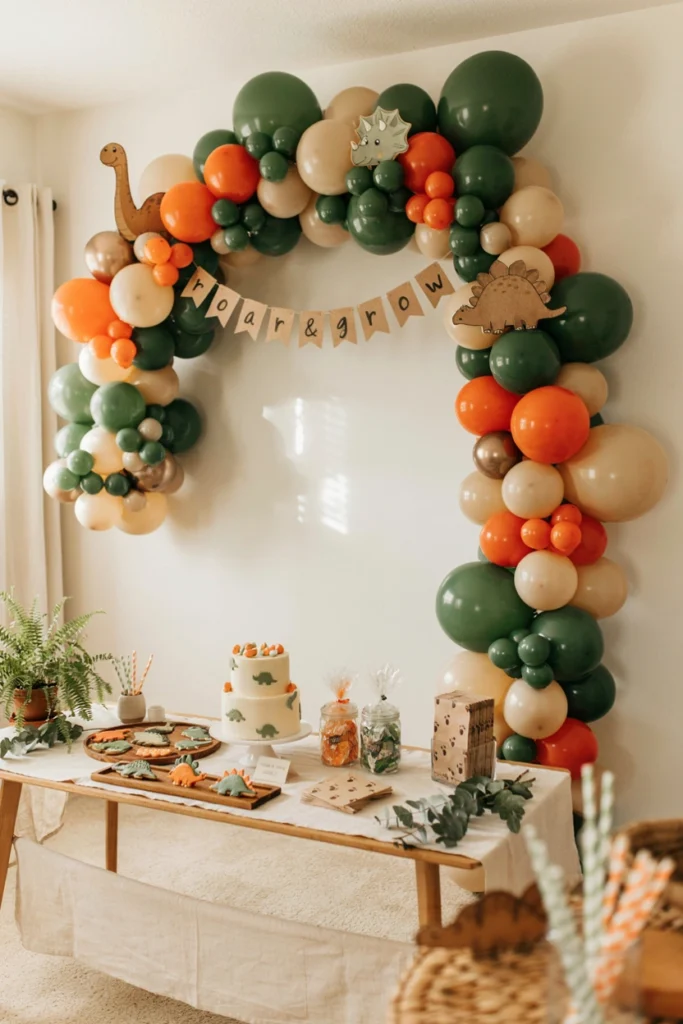 Green and orange balloon arch with small dinosaur cutouts tucked between balloon clusters