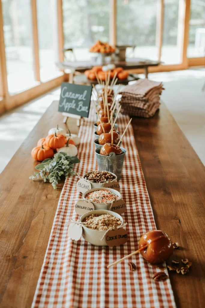 A dessert bar with caramel apples on sticks, bowls of crushed nuts and sprinkles, and small carved pumpkin signage labeling each topping