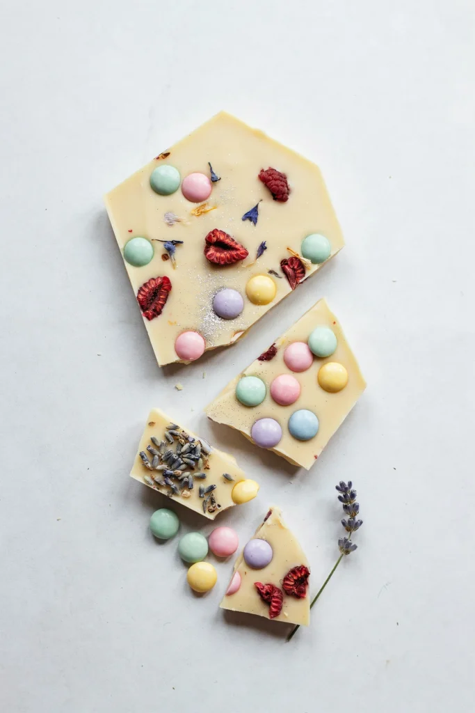 A slab of white chocolate bark broken into irregular pieces. The chocolate is studded with pastel-colored candies, edible glitter, and perhaps dried flowers or freeze-dried berries, creating a crystalline, magical appearance. Pieces are scattered on a white surface.