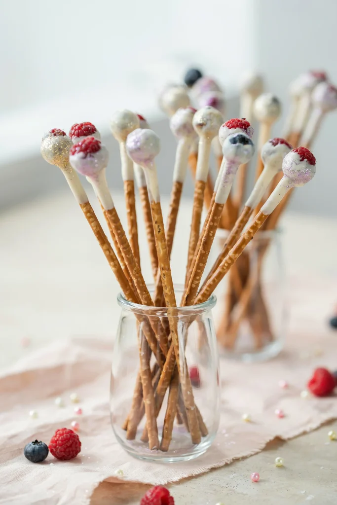 A cluster of pretzel rod "wands" dipped partway in white chocolate and decorated with edible glitter in pastel colors (pearl, pastel pink, lavender). Fresh raspberries and blueberries are attached to the white chocolate end. They're standing upright in a small glass or vase, looking like an actual display of magical wands.