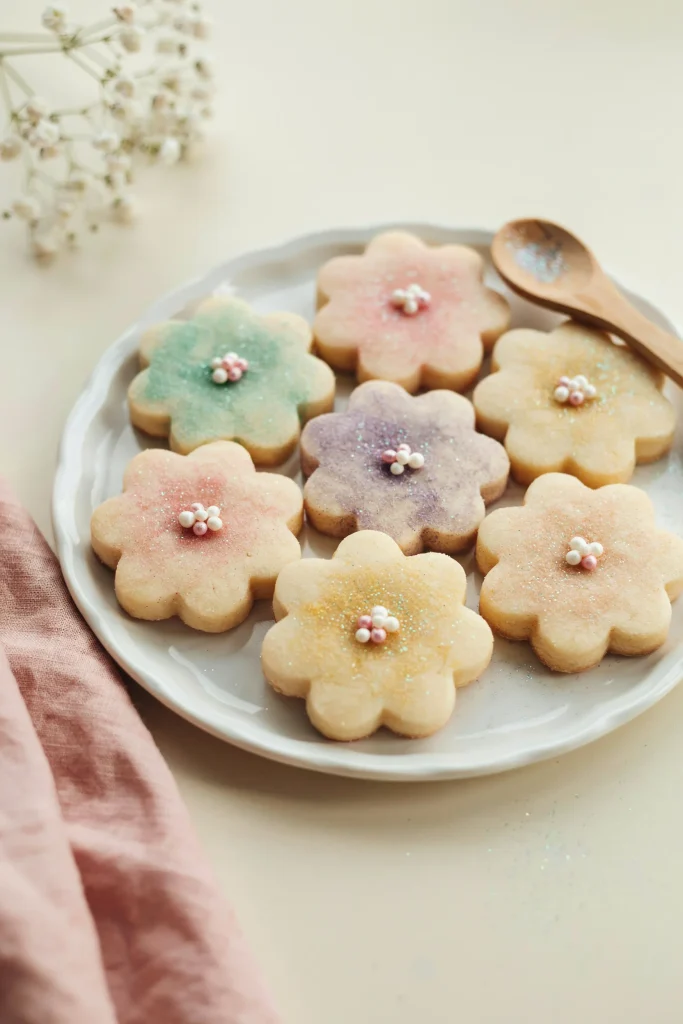 A plate of simple sugar cookies cut into flower shapes (5-petaled or daisy-like). Each cookie is dusted with pastel-colored edible glitter, creating a sparkly, delicate effect. Some cookies have tiny edible pearls in the center.