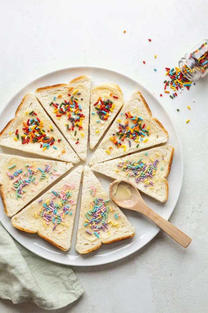 Colorful slices of white bread spread with pale butter and covered in rainbow sprinkles or pastel-colored sprinkles. Crusts trimmed. Cut into triangles or rectangles and arranged tightly on a white plate so the bright colors are fully visible.