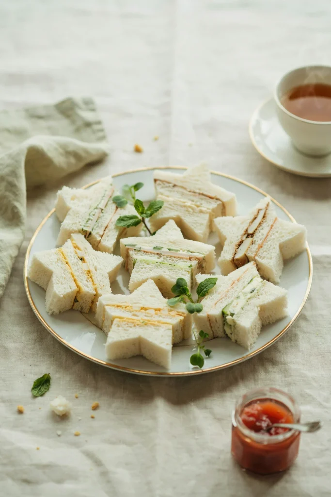 Neat rows of white-bread tea sandwiches cut into five-pointed star shapes. The visible sandwiches show creamy white or pale yellow filling (cucumber & cream cheese, or chicken salad). They're arranged on a white or pale pastel plate with small garnish leaves between them.