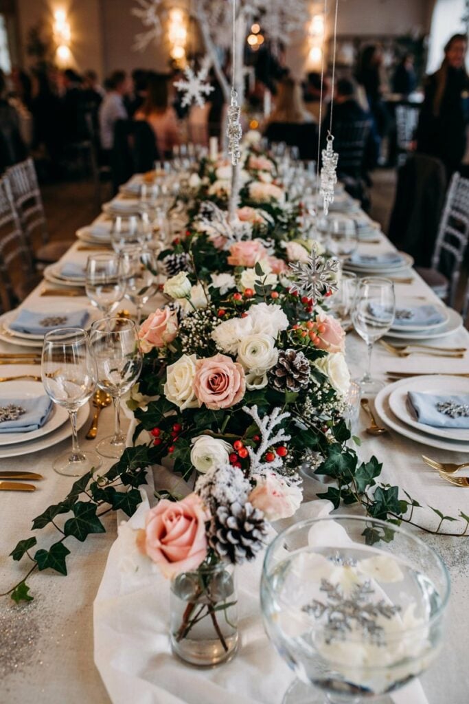 A low, horizontal floral arrangement running down the center of a table. White garland base studded with white flowers, blush roses, baby's breath, and cascading ivy. Candles nestled between flowers. Lush and romantic