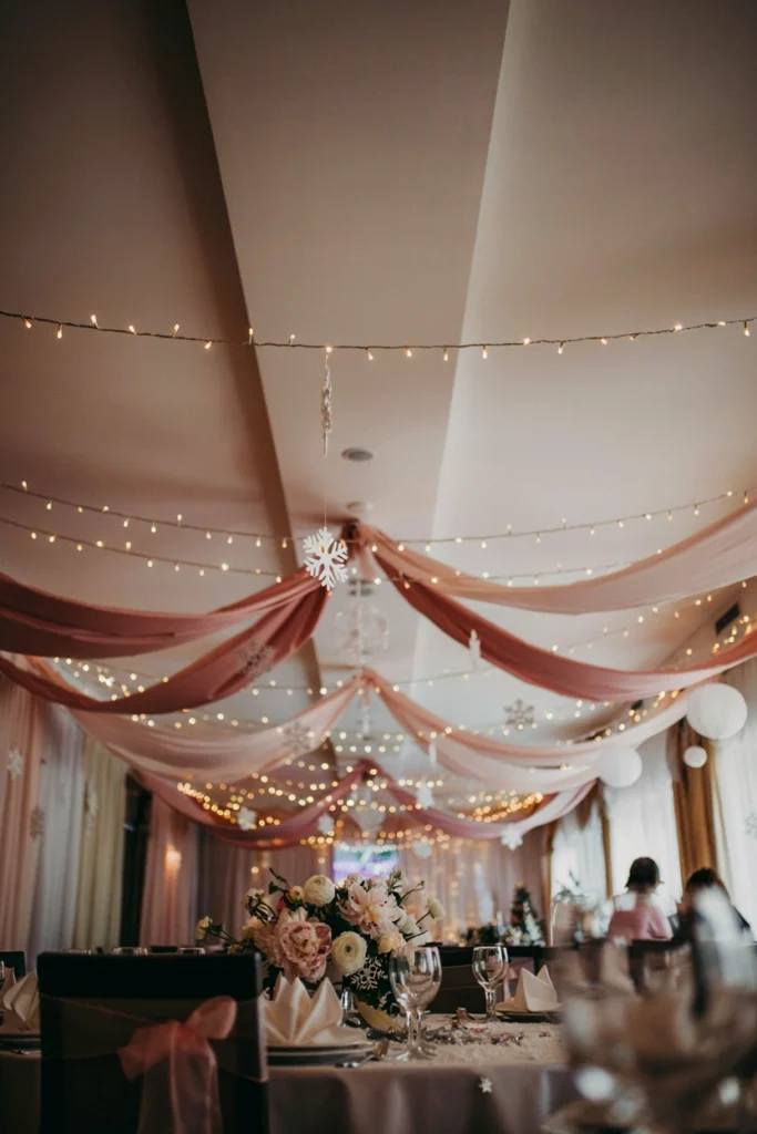 A ceiling strung with overhead fairy lights (warm white, not cold blue), with soft blush tulle or sheer fabric draped between them. Creates a canopy effect. Warm, intimate, magical

