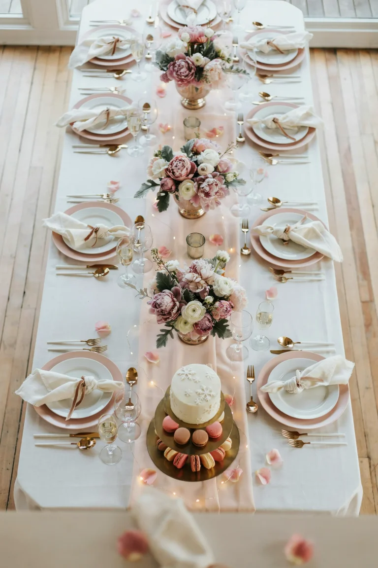 A sweeping overhead shot of a pink and white winter wonderland baby shower setup with fairy lights, frosted florals, white linens, and delicate champagne-colored accents. Soft, dreamy lighting