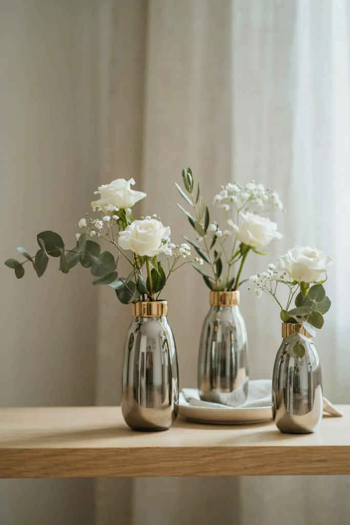 Three smaller vases with sparse, intentional white florals—a few white roses, minimal baby's breath, one greenery stem per vase. Gold-rimmed or mercury gold vases. Modern, sophisticated, Scandinavian-inspired.