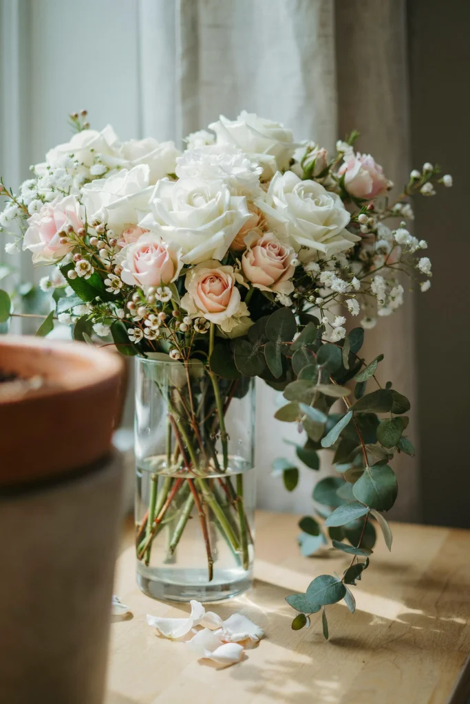 A lush, abundant arrangement of white roses with touches of pale blush roses, baby's breath, and eucalyptus spilling from a tall clear glass vase. Garden-style, romantic, very full and generous.]