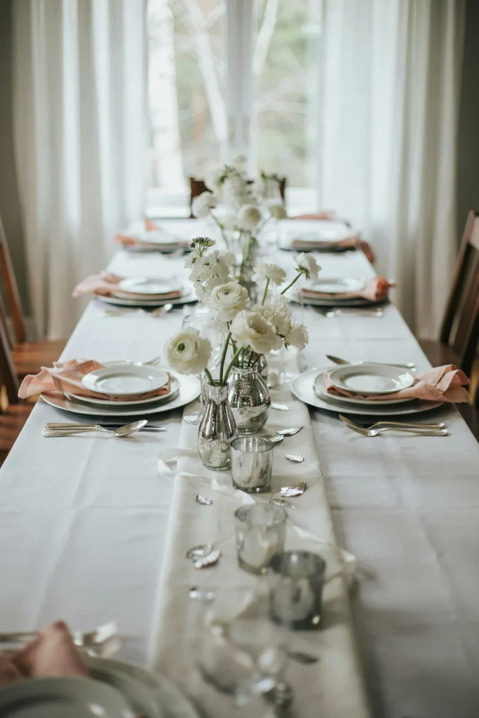 A winter wonderland display featuring pure white flowers, bright white linens, blush pink napkins folded artfully, silver mercury glass vases, and sparkling silver accents throughout. Cool-toned, icy-feeling but still warm