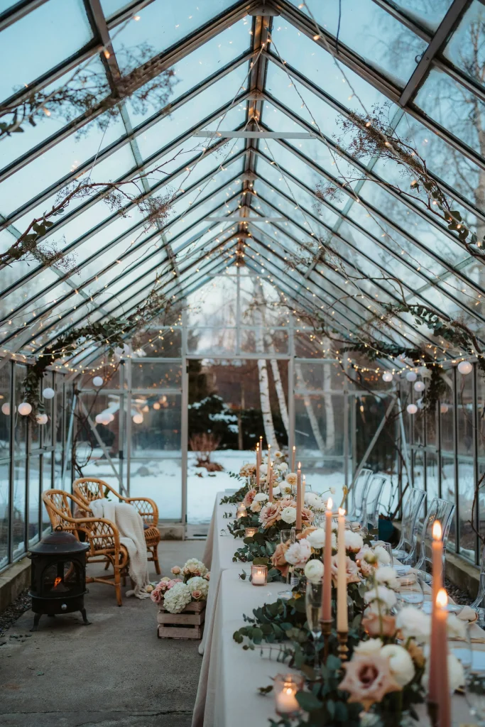 A glass greenhouse or covered outdoor garden space decorated for winter. White and blush flowers, string lights, comfortable seating, heaters or blankets visible. The glass walls are decorated with frosted branches or garland. Magical, weather-protected, unique venue choice.
