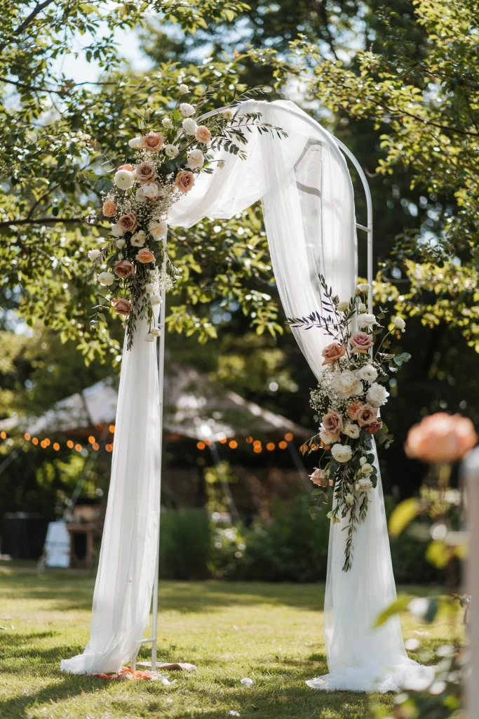 A freestanding or wall-mounted arch draped in flowing white tulle, with white and blush flowers woven throughout. Elegant, romantic, immediately photo-ready. Guests can stand in front for portraits