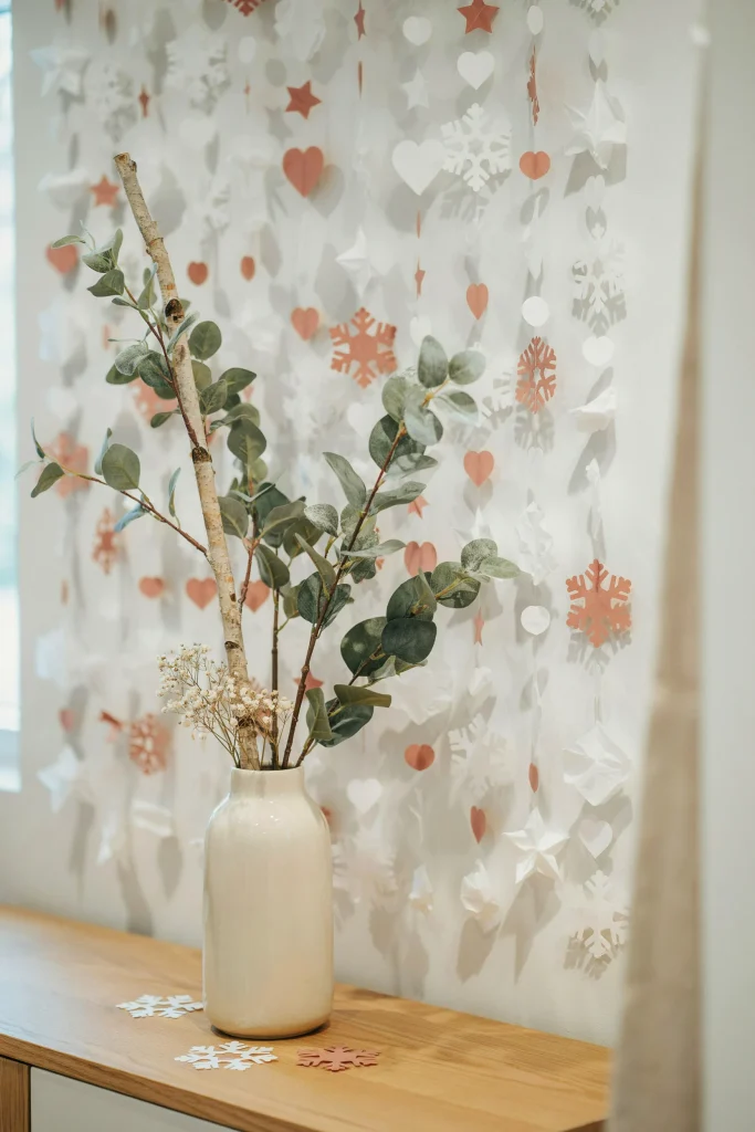 A background wall covered in white and blush tissue paper cutouts (snowflakes, hearts, stars), layered and overlapping. Frosted branches arranged in front in a large vase. Whimsical, dimensional, completely DIY-able