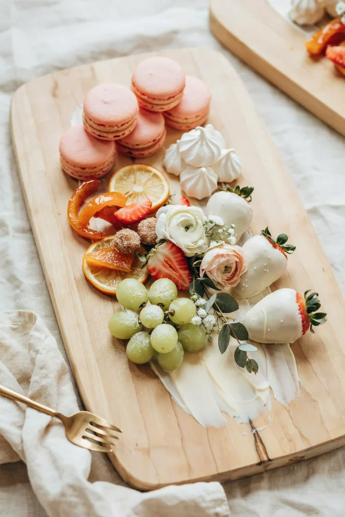 A large wooden board spread with white chocolate, blush macarons, white meringues, candied fruit, white chocolate-covered strawberries, frosted grapes, and a few fresh flowers for garnish. Abundant, generous, totally shareable