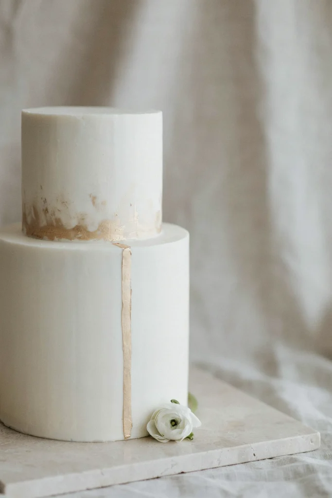 A clean, modern white cake (fondant or perfectly smoothed buttercream) with minimal decoration: a thin gold leaf stripe down one side, or delicate gold dust brushed on. Very high-end, minimalist aesthetic