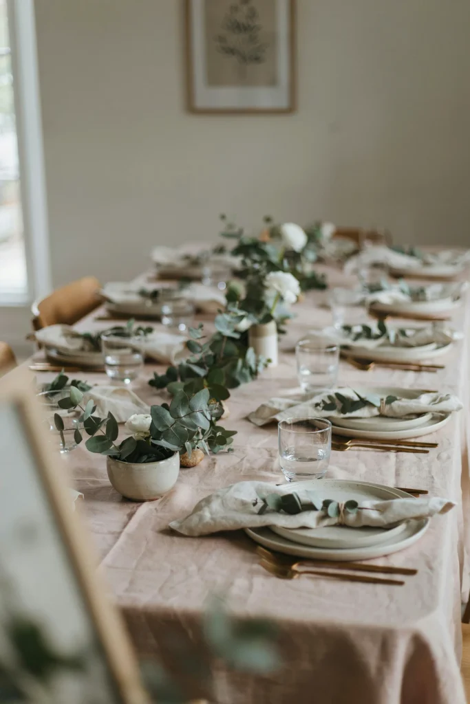 Blush tablecloth with ivory napkins tied loosely with fresh eucalyptus sprigs or a sprig of greenery. Simple, organic, warm. Each place setting shows the napkin detail clearly