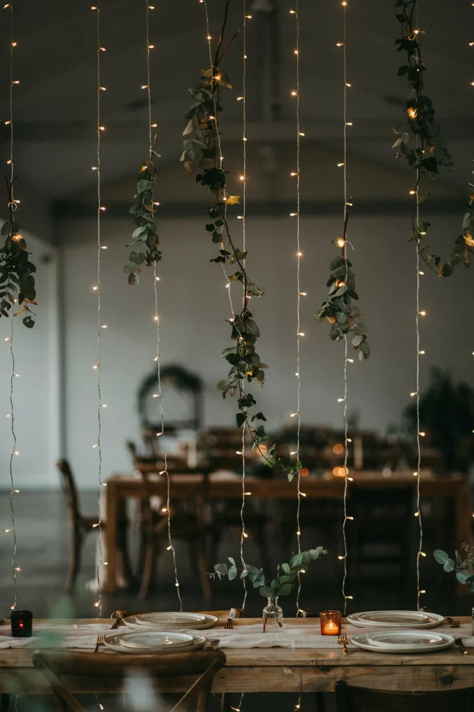 String lights (warm white fairy lights) draped vertically from ceiling to table, with eucalyptus or ivy woven between the strands. Creates a curtain effect of light and greenery. Dramatic, textured, very Pinterest-worthy