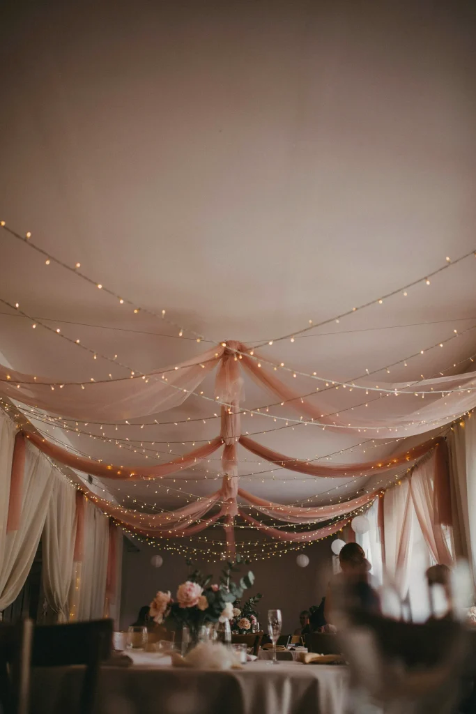 A ceiling strung with overhead fairy lights (warm white, not cold blue), with soft blush tulle or sheer fabric draped between them. Creates a canopy effect. Warm, intimate, magical