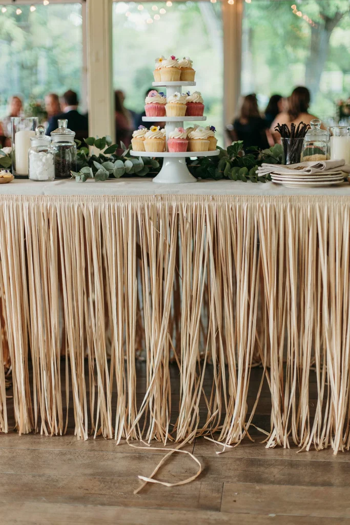  A dessert table skirted in natural raffia-toned grass fringe, cupcake stand on top