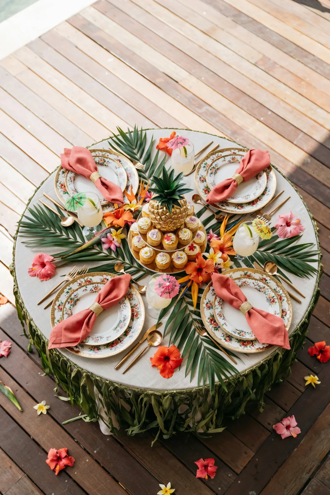 An overhead shot of a tropical party table with a grass table skirt, gold floral tableware, a palm leaf garland, and a pineapple cupcake stand. Bright, sunlit, colorful.