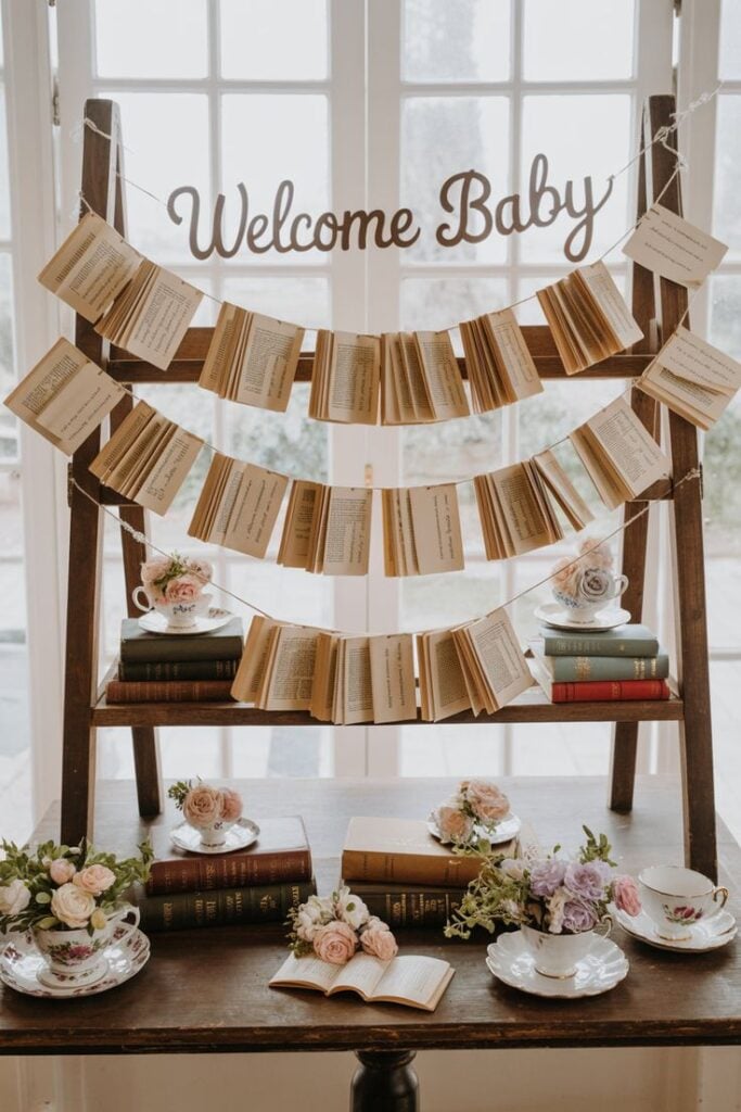 Welcome Baby banner hung above a ladder shelf decorated with hanging book-page garlands, teacups, and stacked antique books