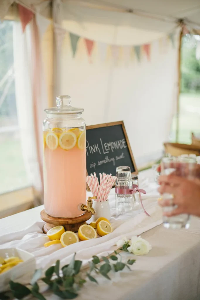 baby shower drink station - A glass beverage dispenser filled with pink lemonade sitting on a wooden stand, surrounded by lemon slices and striped paper straws