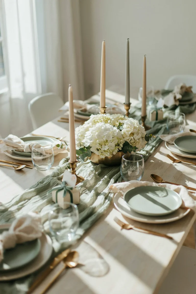 An overhead shot of a beautifully styled baby shower table with a sage cheesecloth runner, a lush ivory hydrangea centerpiece, gold accents, and soft natural light — mood should feel warm, curated, and achievable rather than overly staged.
