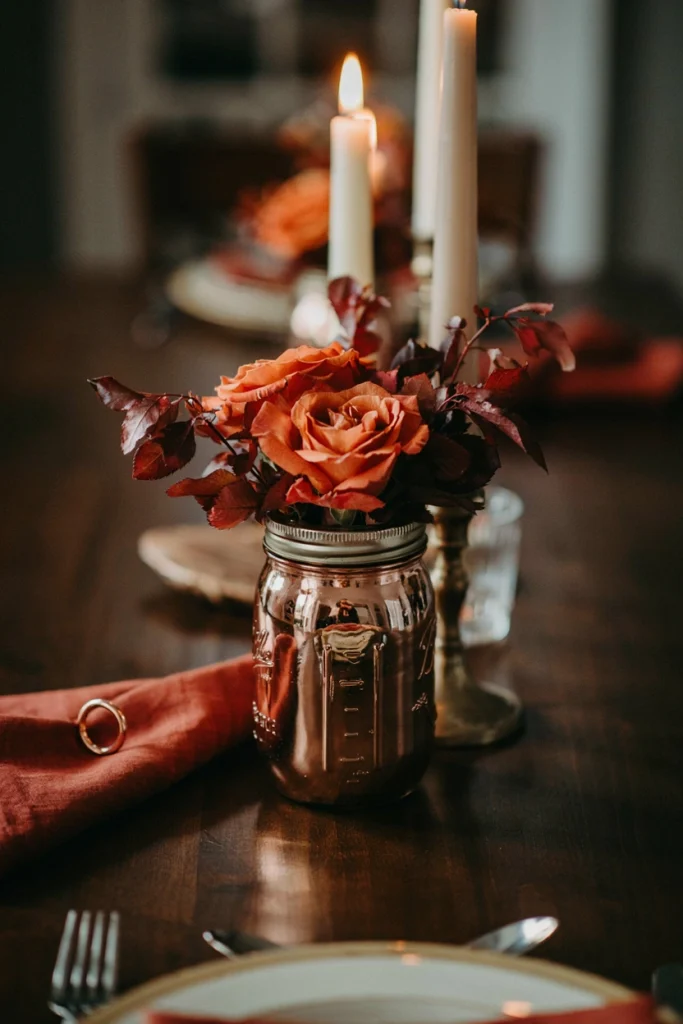 Mason jar painted with metallic copper accents, filled with rust-orange roses and dark red foliage on a candlelit formal table