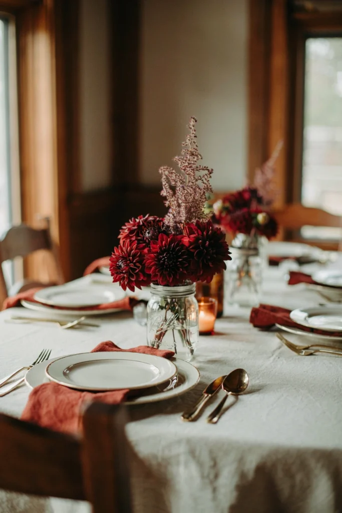 Mason jar filled with deep burgundy dahlias and a single stem of smoke bush on a formally set Thanksgiving table with gold flatware