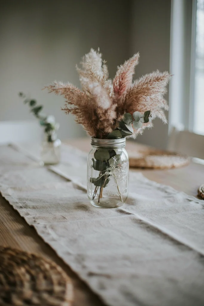 Mason jar filled with fluffy dusty-pink pampas grass and sage-green dried eucalyptus on a linen runner