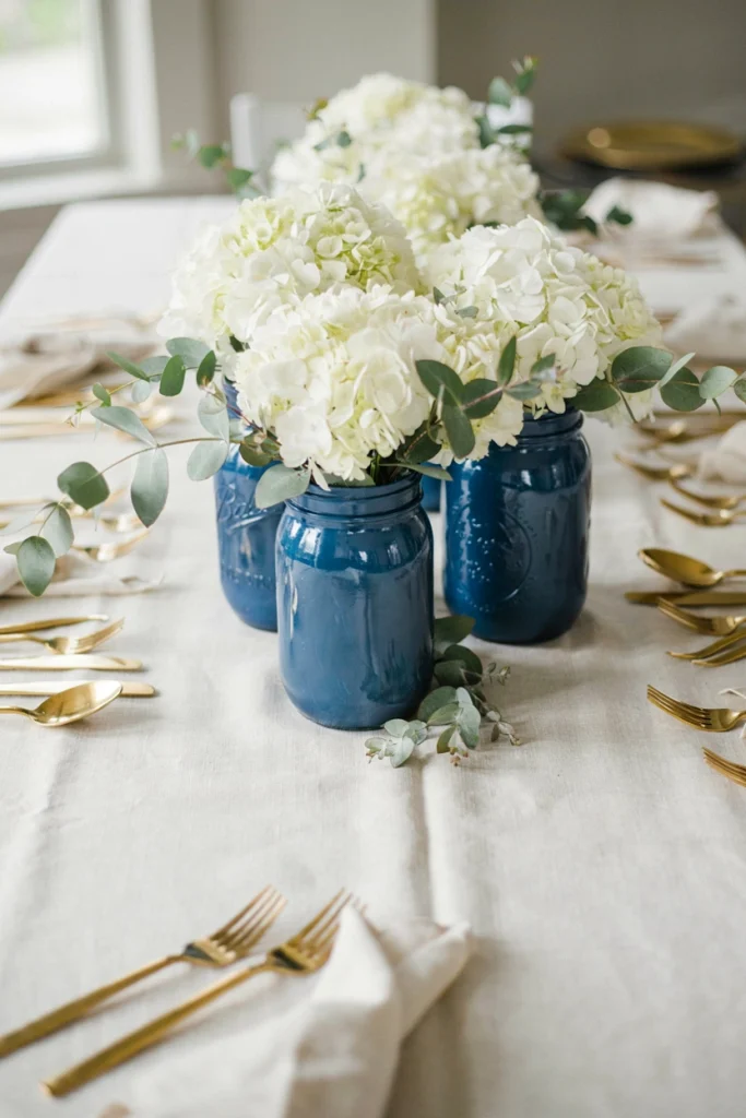 Mason jars painted royal blue and filled with white hydrangeas and silver eucalyptus on a white linen tablecloth with gold flatware