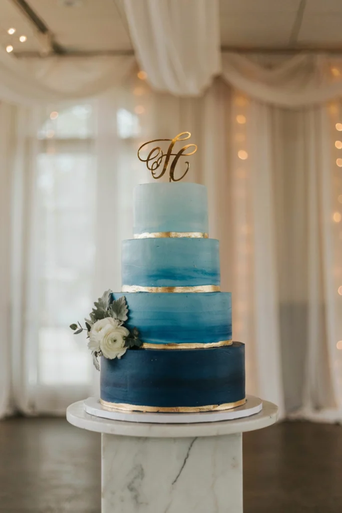 Three-tier cake with buttercream fading from navy to pale sky blue, topped with a gold cake topper